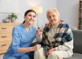 Pflegekraft sitzt lächelnd neben einer älteren Dame im Wohnzimmer